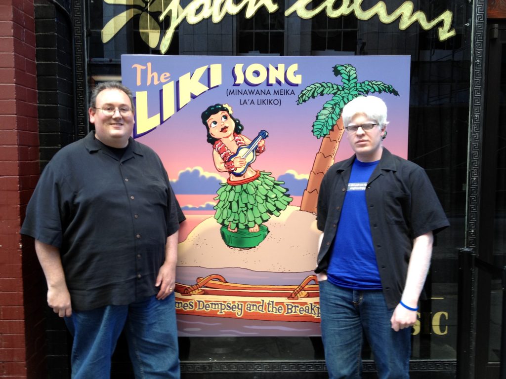 James Dempsey and Darren Minifie standing in front of The Liki Song poster, outside of John Colins bar in San Francisco