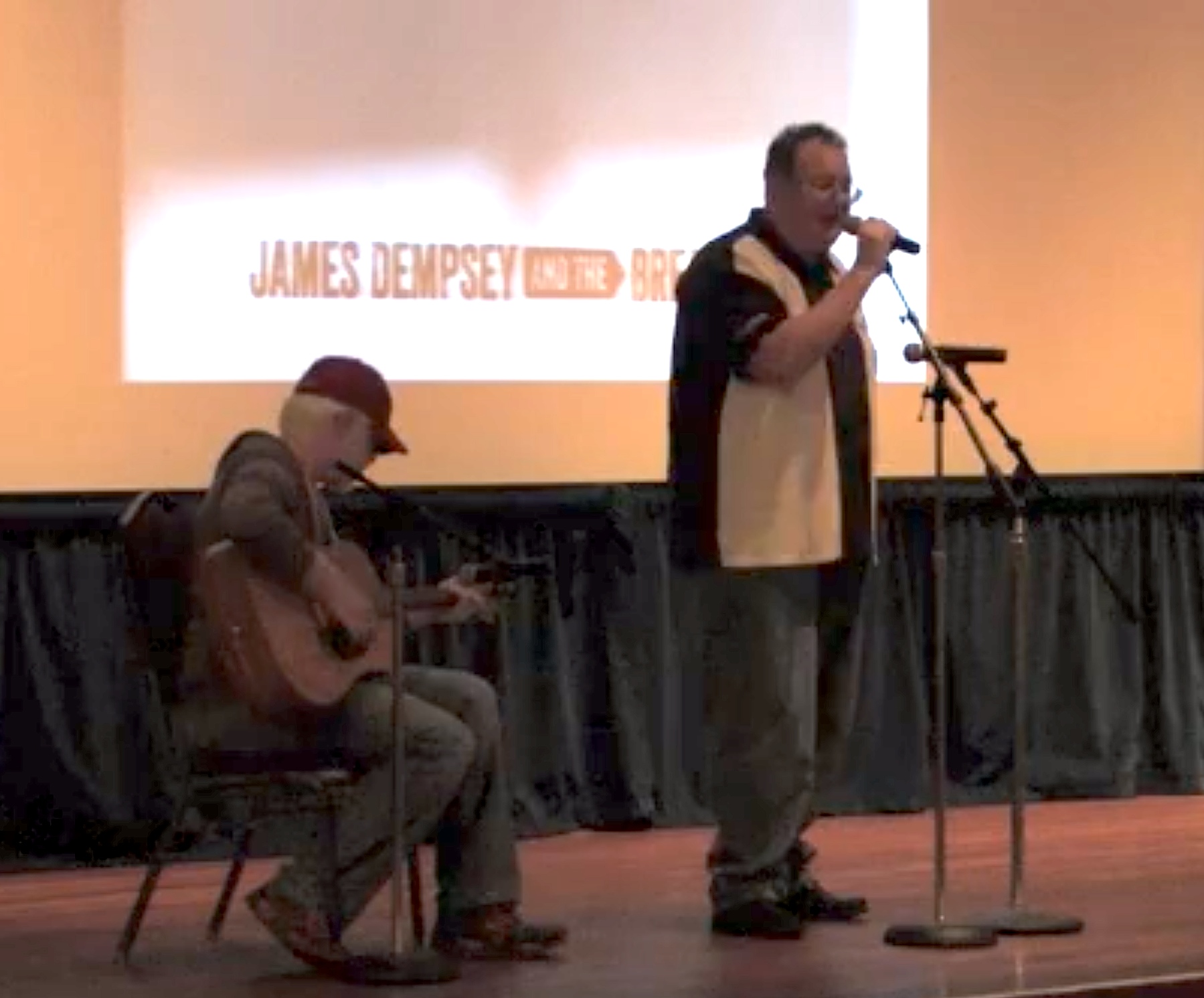James Dempsey singing at a microphone with Darren Minifie sitting playing acoustic guitar.