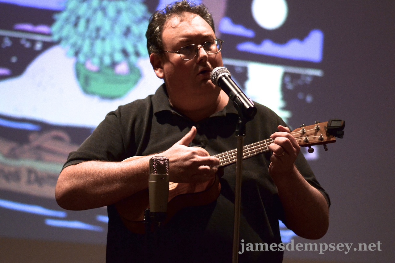 James Dempsey playing ukulele with Liki Song graphic projected in background